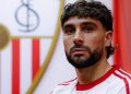 Neal Maupay posando con la camiseta del Sevilla y el escudo al fondo. | Imagen: perfil de X @SevillaFC.