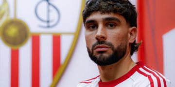 Neal Maupay posando con la camiseta del Sevilla y el escudo al fondo. | Imagen: perfil de X @SevillaFC.